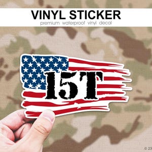 以下が含まれることがあります： アメリカ国旗のデザインと「15T」の文字が入ったビニールステッカー。ステッカーは白い縁取りがあり、迷彩柄の背景に手で持たれています。「VINYL STICKER」の文字が上部にあります。プレミアム防水ビニールデカール。