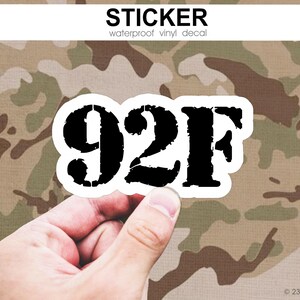 May include: A white-bordered sticker with the black text "92F" is held against a camouflage background. The sticker is labeled "waterproof vinyl decal" at the top. The background has a brown, green, and tan pattern.