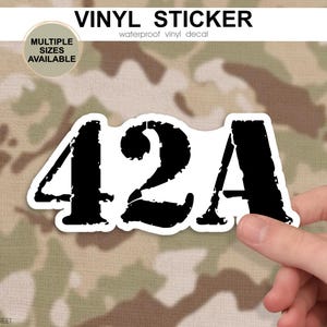 May include: A white vinyl sticker with the number "42A" in a distressed black font. The sticker is held by a hand against a camouflage background. The text "VINYL STICKER" and "waterproof vinyl decal" are at the top.