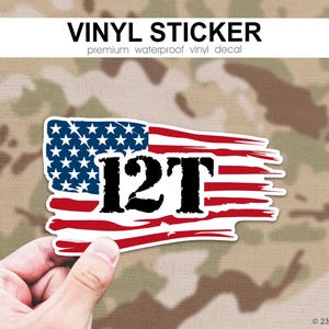 May include: A vinyl sticker with a distressed American flag design and the text "12T" in black. The sticker has a white border and is held against a camouflage background. The text "VINYL STICKER" and "premium waterproof vinyl decal" are at the top.