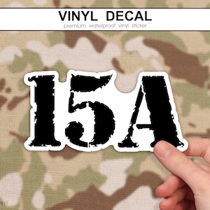 May include: A black and white vinyl decal with the text "15A" in a distressed font. The decal is held by a hand against a camouflage background. The top of the decal reads "VINYL DECAL".