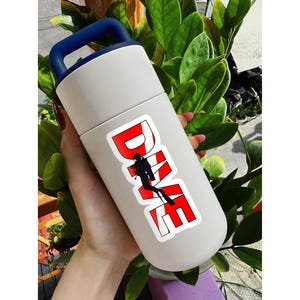 May include: A white insulated water bottle with a dark blue handle on the lid. The bottle features a sticker with the word DIVE in bold red letters, with a black silhouette of a scuba diver integrated into the letter I.