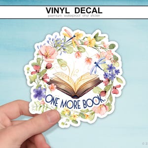 Peut inclure: Un autocollant en vinyle découpé représentant un livre ouvert entouré d'une couronne aquarelle de fleurs roses, jaunes et bleues avec des feuilles vertes et des libellules. Le texte ONE MORE BOOK est imprimé en lettres capitales bleu foncé.