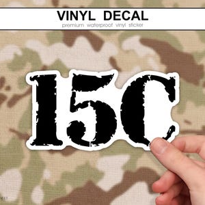May include: A white vinyl sticker featuring the text "15C" in a distressed black font. The sticker is held by a hand against a camouflage background. The top of the image reads "VINYL DECAL" in white text.