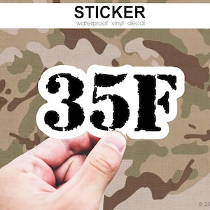 May include: A white sticker with the black text "35F" in a bold, distressed font is held against a camouflage background. The sticker also has the words "STICKER" and "waterproof vinyl decal" in black text.
