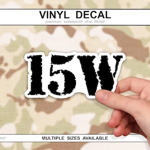 May include: A black and white vinyl decal with the text "15W" in a distressed font. The decal is held by a hand against a camouflage background. The top of the image reads "VINYL DECAL".