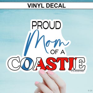 May include: Vinyl decal with the text "PROUD Mom OF A COASTIE" in black, blue, and red. The word "Mom" is in a cursive font. The decal is white with a black outline and is held by a hand with pink nail polish.