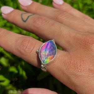 May include: A silver ring with a large, iridescent, teardrop-shaped stone. The stone displays a spectrum of colors, including purple, blue, and green. The ring is set on a hand with manicured nails, against a backdrop of green grass.