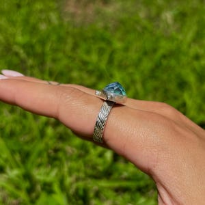 May include: A silver ring with a large, faceted, blue-green gemstone. The ring band features an engraved pattern. The ring is displayed on a hand against a blurred green background.