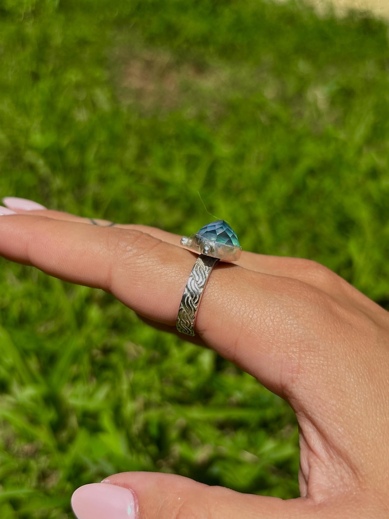 May include: A silver ring with a large, faceted, blue-green gemstone. The ring band features an engraved pattern. The ring is displayed on a hand against a blurred green background.