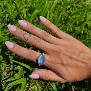May include: A silver ring with a large, teardrop-shaped, iridescent blue and purple gemstone. The ring is set against a backdrop of green grass, showcasing the ring's vibrant colors and design.