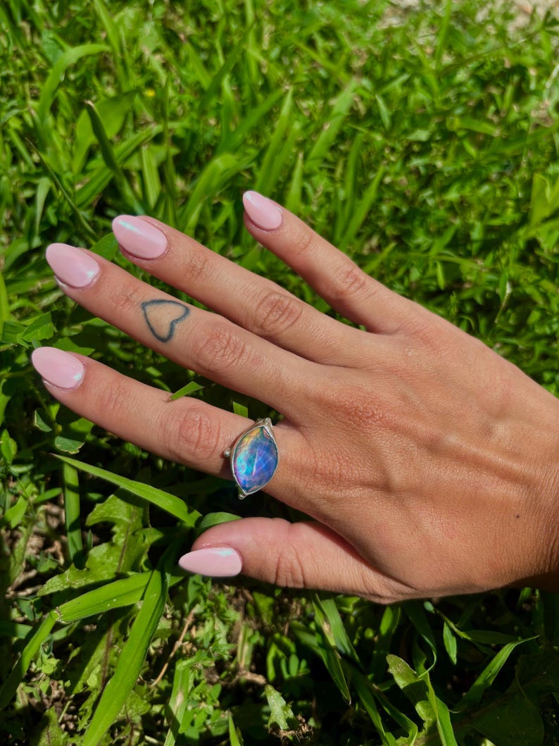 May include: A silver ring with a large, teardrop-shaped, iridescent blue and purple gemstone. The ring is set against a backdrop of green grass, showcasing the ring's vibrant colors and design.