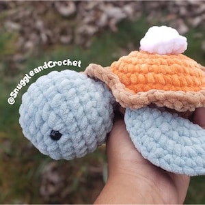 May include: A handmade crochet turtle toy with a light blue body, orange shell, and a white pom-pom on top. The turtle has a black eye and is made of soft, textured yarn. The text "@SnuggleandCrochet" is visible.