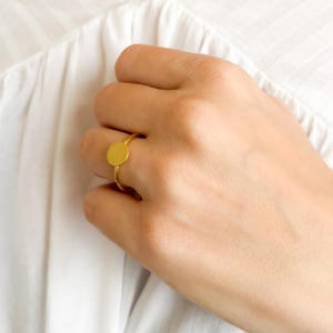 Op de afbeelding: Een goudkleurige ring met een klein, cirkelvormig ontwerp. De ring heeft een dunne band en een plat, rond middelpunt. De ring wordt getoond op een hand, tegen een witte achtergrond. Het ontwerp van de ring is eenvoudig en elegant.