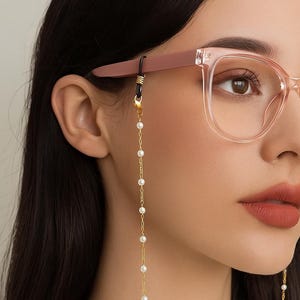 May include: A gold-colored eyeglass chain with white pearls. The chain is attached to the glasses and hangs down, creating a stylish accessory. The glasses are a light pink color.