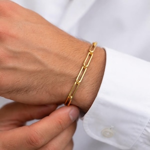 May include: A gold-colored link bracelet is displayed on a wrist, set against a white shirt. The bracelet features a series of elongated, rectangular links. The image is well-lit, highlighting the jewelry's metallic sheen.