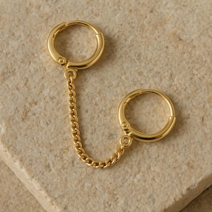 May include: Gold-colored double hoop earring with a chain. The earring features two circular hoops connected by a delicate chain. The hoops have a hinged closure. The jewelry is displayed on a light beige stone surface.