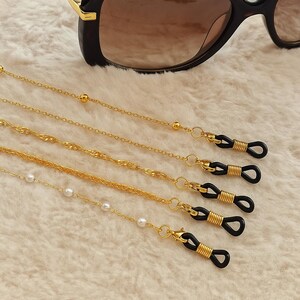May include: Gold-colored eyeglass chains with various chain styles and black rubber loops. One chain features small white pearls. Black sunglasses with brown lenses are in the background. The chains are on a soft, white, textured surface.