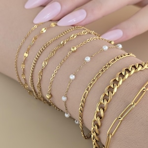 May include: A set of seven gold chain bracelets, including a delicate chain with small pearls, a thicker curb chain, and a chain with small round beads. The bracelets are stacked on a person's wrist.