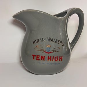 Könnte beinhalten: Graue Keramikkrug mit gebogenem Griff und Ausguss. Der Krug trägt den Text "HIRAM WALKER'S TEN HIGH A TRUE BOURBON" in roter und goldener Schrift. Der Krug hat eine glatte, glänzende Oberfläche.