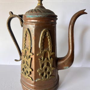 May include: Antique copper and brass coffee pot with ornate brass detailing. The pot features a long, curved spout, a handle, and a decorative lid with a finial. The copper has a warm, aged patina, and the brass accents add a touch of elegance.