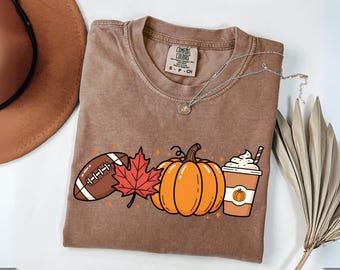 May include: A brown Comfort Colors t-shirt featuring a fall-themed graphic. The design includes a football, a red maple leaf, an orange pumpkin, and a coffee cup with whipped cream. A silver necklace with a pendant is also visible.