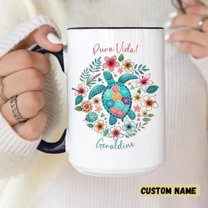 May include: White ceramic mug with a black handle and rim. The mug features a colorful sea turtle design surrounded by flowers and the words "Pura Vida!" and "Geraldine". The bottom right corner has the text "CUSTOM NAME".
