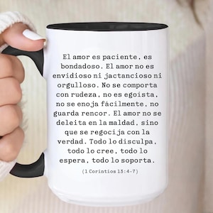 May include: White ceramic mug with a black handle and rim, featuring Spanish text from 1 Corinthians 13:4-7. The mug is held by a person wearing a white sweater. The text describes love's characteristics.