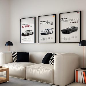May include: Three framed posters of sports cars, each with a different car model and brand. The posters are hung on a white wall above a white couch with black and white patterned pillows. The posters feature a white Nissan GT-R, a white Porsche 911 GT3 RS, and a black Ford Mustang Mach-1.
