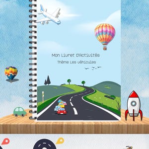 Peut inclure: Un livre d'activités relié en spirale bleu et blanc avec le titre "Mon Livret D'activités" et le sous-titre "Thème Les Véhicules". La couverture présente un avion de dessin animé, un ballon à air chaud et des voitures qui roulent sur une route sinueuse.
