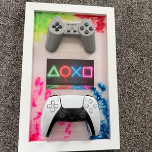 May include: A white rectangular frame containing a gray Sony PlayStation controller at the top and a white and black PlayStation 5 controller at the bottom. Between the controllers is a rectangular print featuring neon green, red, blue, and pink symbols.