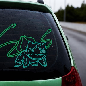 May include: A green car with a green and blue outline sticker of a Pokemon character on the back window. The character has a long tail and a large head with a wide mouth.