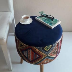 May include: A blue velvet and multi-colored patterned ottoman with wooden legs. A book and a cup of coffee are on top of the ottoman.