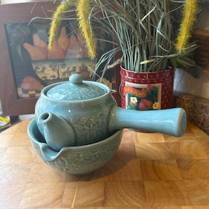 May include: A light blue ceramic teapot with a matching saucer. The teapot has a curved handle and a lid with a small knob. The set is placed on a wooden surface. Decorative floral patterns are carved into the teapot and saucer.