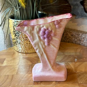 Peut inclure: Un vase en céramique rose avec une base carrée et un dessus évasé. Le vase présente un motif de grappe de raisin en relief en violet et crème. Le vase est exposé sur une surface en bois.