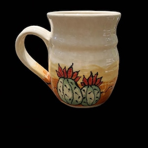 May include: Cream-colored ceramic mug with a hand-painted desert scene. The mug features two green cacti with red flowers against a backdrop of tan and orange hues. The handle is also cream-colored, and the mug has a slightly textured surface.