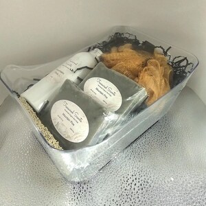 May include: A clear plastic gift basket filled with bath products. The basket contains a white bottle, two dark green wrapped soaps, and a tan loofah. The soaps have white labels that read "Treasured Scents Mountain Pine."