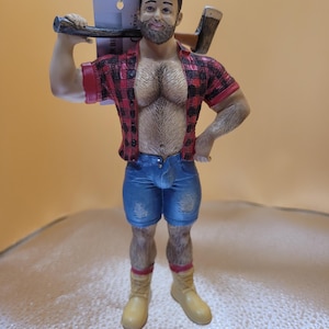 May include: A decorative ornament of a muscular figure with a beard, wearing a red and black plaid shirt, blue denim shorts, and yellow boots. The figure is holding an axe over its shoulder. The ornament has a tag hanging from the top.