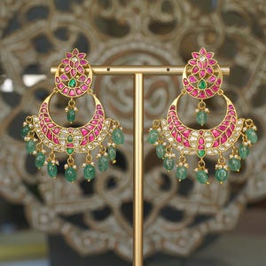 May include: Gold crescent earrings with pink and white detailing, green emerald drops, and floral designs. The earrings are presented on a gold stand against a blurred backdrop.