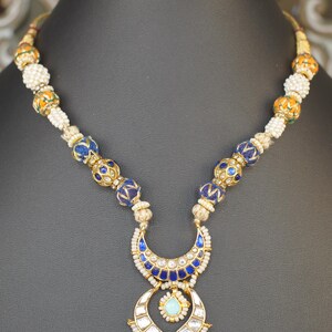 May include: An ornate necklace with a gold chain and pendant. The necklace features blue and gold beads, a crescent-shaped pendant with blue enamel, and pink gemstone drops. The necklace is displayed on a black mannequin.
