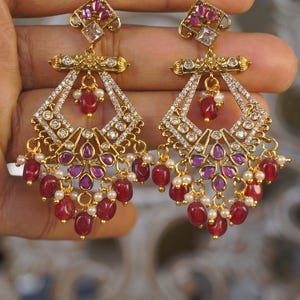 May include: A pair of gold-toned chandelier earrings. Each earring features a square top with pink gemstones and a clear gemstone. The earrings have a diamond-shaped design with clear gemstones and dangling red beads and small white pearls.