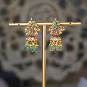 May include: Gold earrings with detailed design. Each earring has a floral pattern with green and red gemstones, and a cluster of green beads. The earrings are displayed on a gold stand.