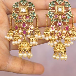 May include: A pair of gold-toned dangle earrings featuring a square central stone, green and pink accents, and multiple small bell-shaped charms hanging from the bottom. Each charm is adorned with tiny white beads, creating a layered design.