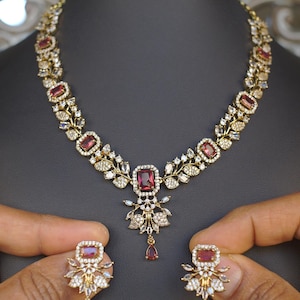 May include: A gold-toned necklace and earring set featuring ruby-colored rectangular and teardrop-shaped gemstones. The necklace and earrings are adorned with smaller clear gemstones and floral designs. The jewelry is displayed on a black mannequin head.