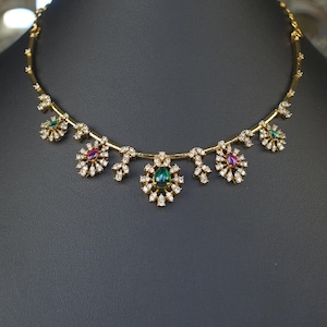 May include: A gold-toned necklace featuring a series of floral-inspired pendants set with clear white stones. The central pendant showcases a teardrop-shaped green stone, while other pendants feature alternating teardrop-shaped green and pink stones.