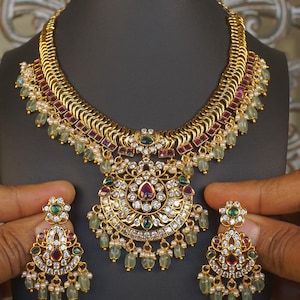 May include: A gold-toned necklace and matching earring set featuring intricate metalwork, embedded with clear white stones, deep red rectangular accents, and small green teardrop-shaped beads. The central pendant is circular with floral motifs.
