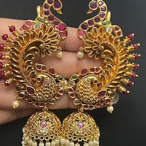 May include: A pair of gold-toned jhumka earrings featuring intricate peacock motifs adorned with small red stones, clear crystals, and green accents. The bell-shaped base is embellished with dangling white pearls and fine metal filigree detailing.