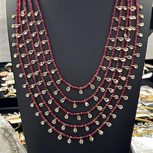May include: A multi-strand necklace featuring five rows of small, deep red faceted beads. Each strand is accented with evenly spaced, clear, teardrop-shaped stones set in gold-toned metal, creating a cascading, layered design on a black display.