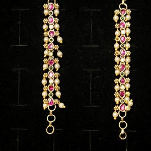 May include: A pair of gold-tone linear earrings featuring a vertical arrangement of teardrop-shaped pink stones and small white pearls. The design includes intricate metal beadwork accents and a small circular link at the bottom of each earring.