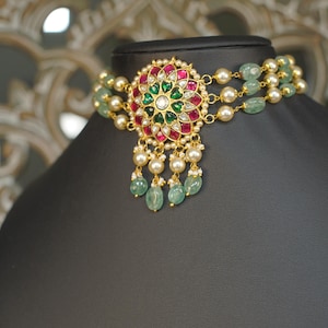 May include: A gold-toned choker necklace with a central floral pendant. The pendant features red, green, and white gemstones. The necklace has multiple strands of pearls and green beads, with dangling green beads below the pendant.